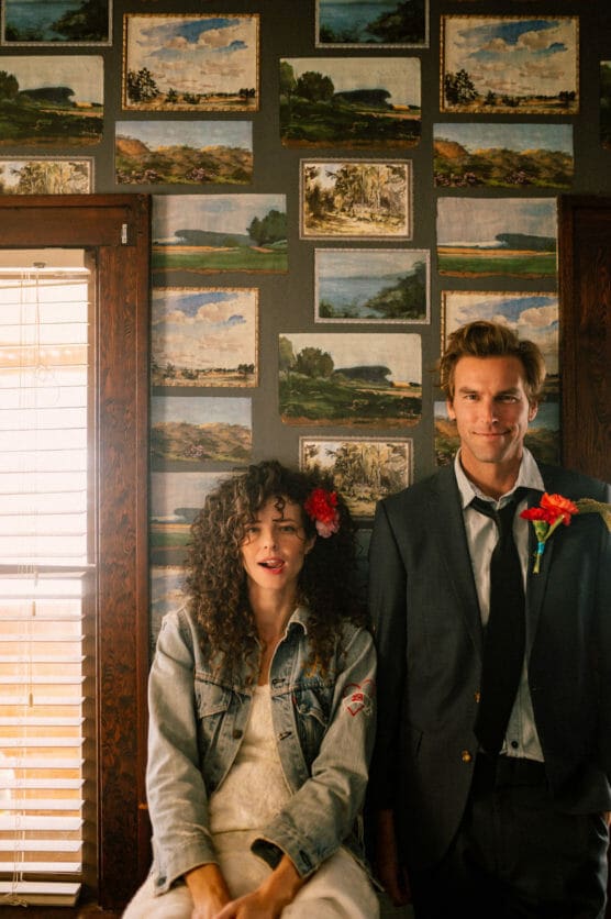 A woman with curly hair and a red flower in her hair, wearing a denim jacket, sits beside a man in a suit with a red flower on his lapel. They pose in front of a wall decorated with landscape paintings. by Justin Salem Meyer