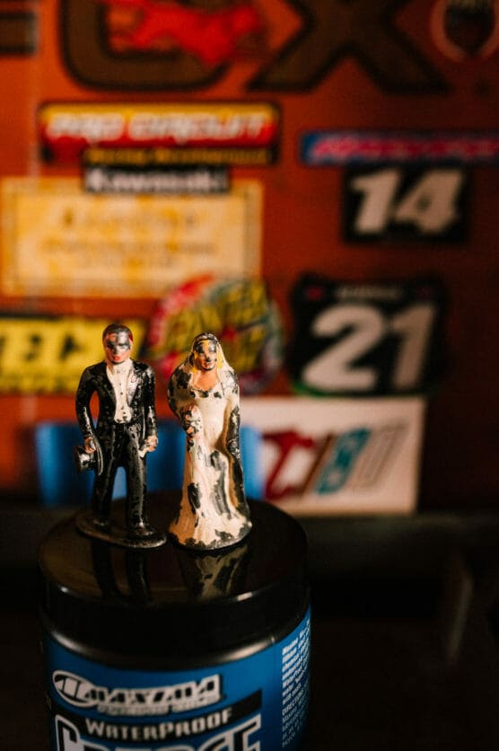 Two small bride and groom figurines stand on top of a blue container, with a blurred background featuring colorful racing number stickers and logos on a wall. by Justin Salem Meyer