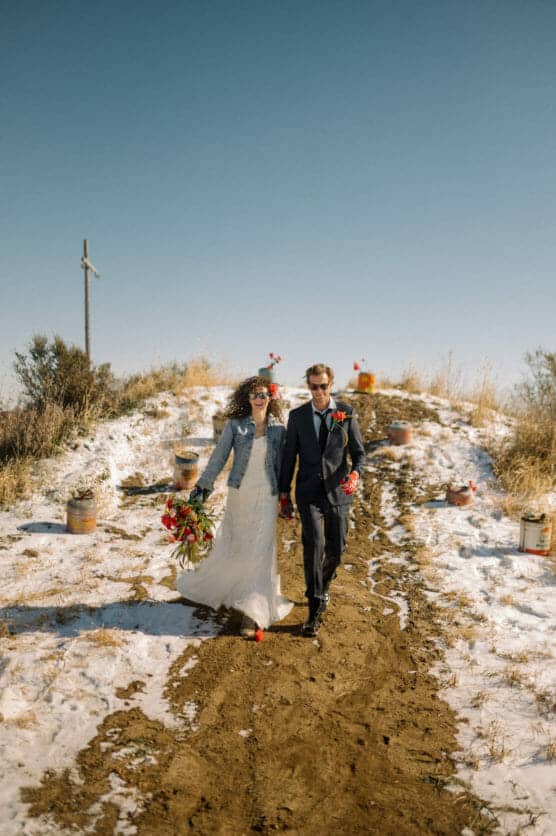 A bride in a white dress and denim jacket, and a groom in a dark suit, walk hand-in-hand down a dirt path with patches of snow, under a clear blue sky, surrounded by dry grass and colorful flowers. by Justin Salem Meyer