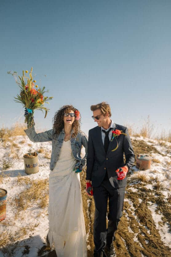 A smiling bride in a white dress and denim jacket holds a bouquet up while walking with a groom in a dark suit and sunglasses on a snowy, grassy hillside. Both look joyful and relaxed under a clear blue sky. by Justin Salem Meyer