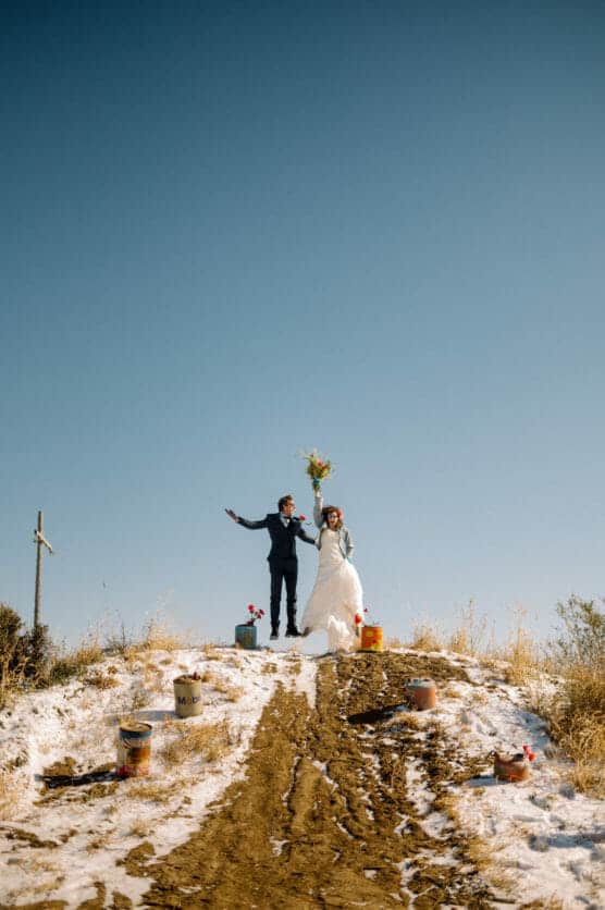 A newlywed couple stands on a snowy hilltop under a clear blue sky, the bride holding a bouquet up high. The groom stands beside her with arms outstretched. Paint cans are scattered on the slope leading up to them. by Justin Salem Meyer
