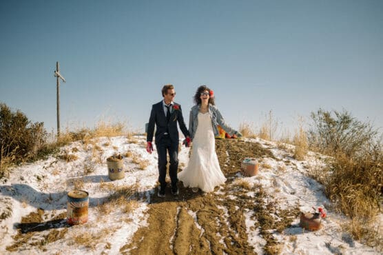 A bride and groom in wedding attire walk hand in hand on a snowy, grassy hill under a clear sky, both wearing sunglasses and red gloves, with scattered paint cans and a wooden cross nearby. by Justin Salem Meyer
