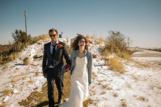 A bride and groom walk hand-in-hand outdoors on a snowy path, both smiling and wearing sunglasses. The bride has curly hair and wears a white dress; the groom wears a suit with a red flower boutonniere. The scene is sunny with a clear sky. by Justin Salem Meyer