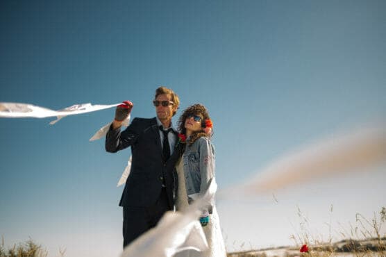 A couple dressed in quirky formal and casual attire stands outdoors under a clear blue sky, holding long white ribbons that flutter in the wind. Both wear sunglasses, and the scene has a playful, unconventional vibe. by Justin Salem Meyer