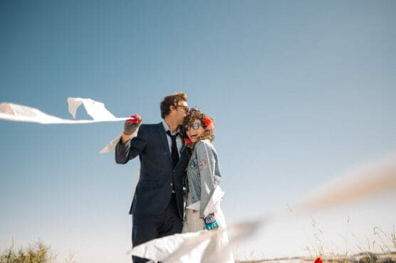 A man in a suit kisses a smiling woman wearing sunglasses and a denim jacket, with white streamers flowing in the wind against a clear blue sky. They appear joyful outdoors. by Justin Salem Meyer