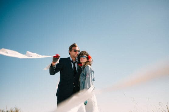 A man in a suit and a woman in a denim jacket, both wearing sunglasses, stand together outdoors under a clear blue sky with white streamers blowing in the wind. by Justin Salem Meyer