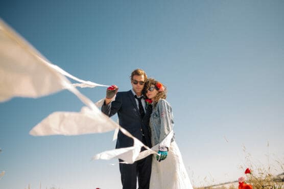 A couple stands outdoors under a clear blue sky. The person on the left wears a suit and sunglasses, while the person on the right wears a white dress, denim jacket, red flower crown, and sunglasses. White flags or streamers blow in the wind. by Justin Salem Meyer