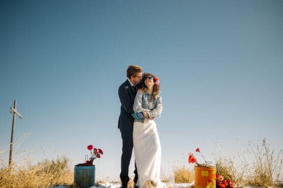 A smiling couple, dressed in wedding attire, stands outdoors on dry grass under a clear blue sky. The woman wears a white dress and flower crown; the man hugs her from behind. Colorful flowers in buckets decorate the scene. by Justin Salem Meyer
