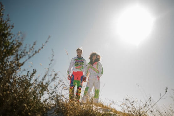 Two people in colorful, neon motocross outfits stand on a grassy hill under a bright, sunny sky. Tall grass surrounds them and the sun shines intensely in the background. by Justin Salem Meyer