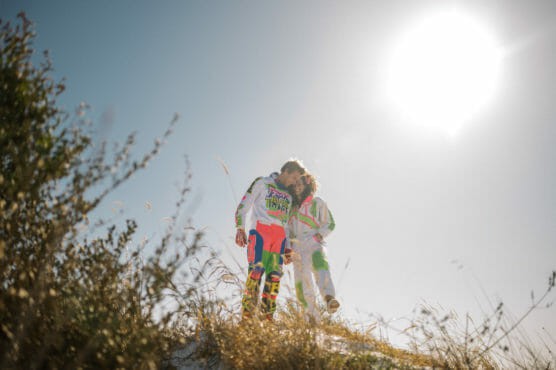 Two people in colorful motocross outfits stand close together on a grassy hill under a bright sun and clear sky, with tall plants in the foreground. by Justin Salem Meyer