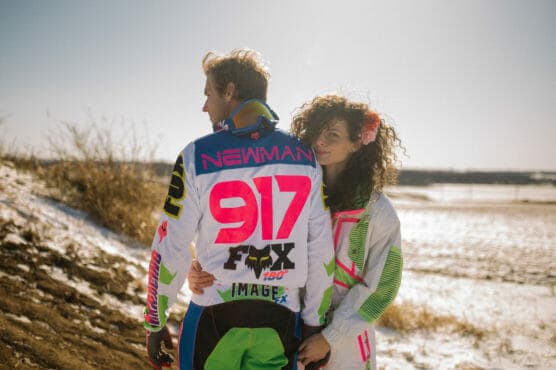 A woman stands outdoors in a snowy field, hugging a man wearing a colorful motocross jacket with NEWMAN 917 FOX on the back. Both wear bright, sporty outfits, and the sun is shining. by Justin Salem Meyer