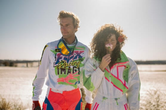 Two people wearing colorful, retro motocross outfits stand outdoors. The man looks to the side, while the woman with curly hair holds a pink flower to her face. The sun is bright, and a sandy landscape is in the background. by Justin Salem Meyer