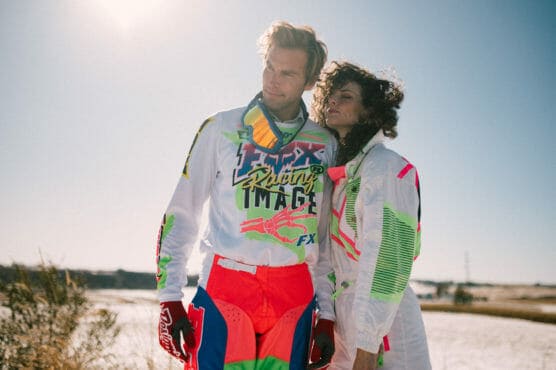 Two people in colorful motocross racing outfits stand closely together outdoors on a sunny day, with one leaning affectionately on the other. The background features a bright sky and natural landscape. by Justin Salem Meyer
