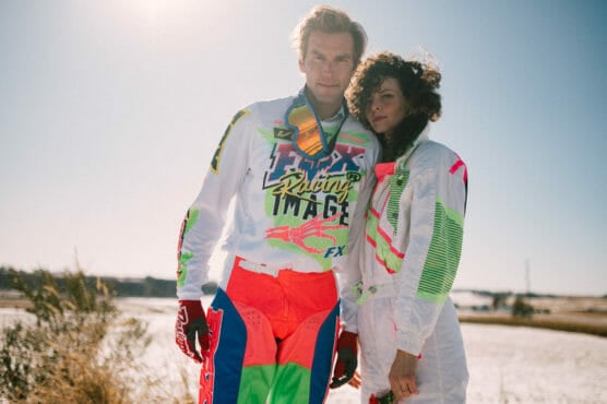 Two people stand outdoors near a body of water, both wearing colorful motocross racing outfits with neon and bright patterns. The sky is clear and sunny, and they pose together, looking at the camera. by Justin Salem Meyer