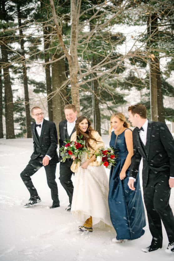 Five people dressed in formal attire walk together in the snow, smiling and holding bouquets. Evergreen trees are in the background, and snow is falling, creating a festive and wintry atmosphere. by Justin Salem Meyer