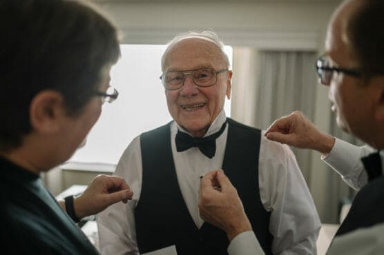 An older man in a bow tie and vest smiles warmly while two people help adjust his outfit in a softly lit room. by Justin Salem Meyer