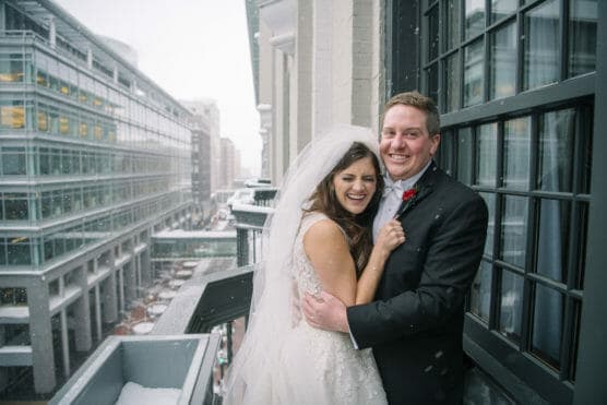 A bride and groom smile and embrace on a snowy balcony outside a city building. The bride wears a white dress and veil; the groom is in a black tuxedo with a red boutonniere. Snow falls softly around them. by Justin Salem Meyer