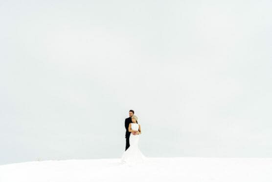 A couple in wedding attire stands embracing on a snowy landscape under a bright, overcast sky, with minimal background elements, creating a serene and minimalist scene. by Justin Salem Meyer