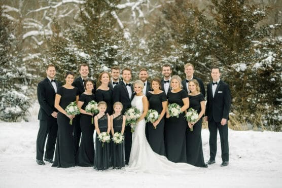 A wedding party poses outdoors in the snow. The bride in white stands with bridesmaids in black dresses and groomsmen in black suits. Three young girls in black dresses hold bouquets. Trees dusted with snow are in the background. by Justin Salem Meyer