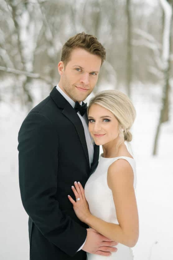 A couple poses closely together outdoors in the snow; the man wears a black tuxedo and the woman wears a sleeveless white dress, showing her engagement ring. Bare trees and a snowy landscape are in the background. by Justin Salem Meyer