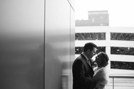 A couple stands close together on a balcony in front of a modern building, gazing into each others eyes. Snow falls softly in the background, and the scene is captured in black and white. by Justin Salem Meyer