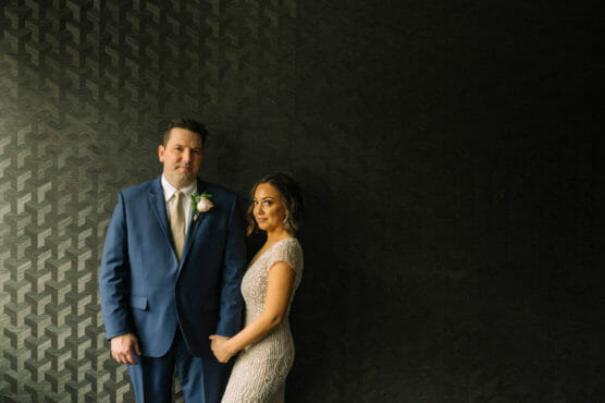 A man in a blue suit with a boutonniere stands beside a woman in a lace dress, holding hands. They are positioned against a dark, patterned wall with ample negative space on the right. by Justin Salem Meyer