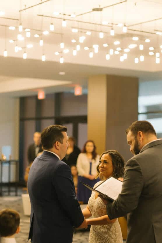 A couple stands facing each other, holding hands, while a man officiates their wedding ceremony indoors under soft, modern lighting. Guests are visible in the background, watching the event. by Justin Salem Meyer