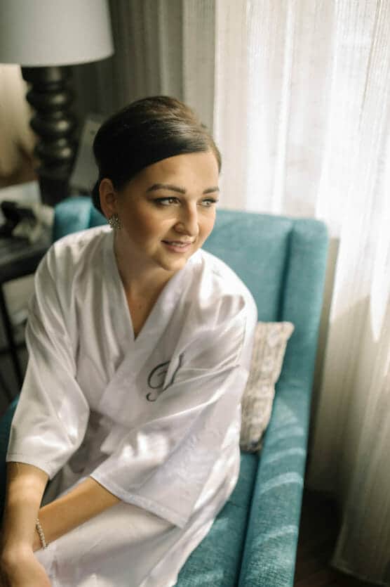 A woman with dark hair in an updo, wearing a white robe, sits on a teal armchair beside a window with sheer curtains, looking out with a gentle smile. by Justin Salem Meyer