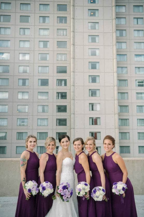 A bride in a white dress stands with five bridesmaids in matching purple dresses, each holding a bouquet of white and purple flowers. They pose outdoors in front of a tall, modern building with many windows. by Justin Salem Meyer