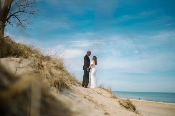 A couple stands on a sandy dune near tall grass, holding hands and gazing at each other under a blue sky with wispy clouds, with a calm beach and water in the background. by Justin Salem Meyer