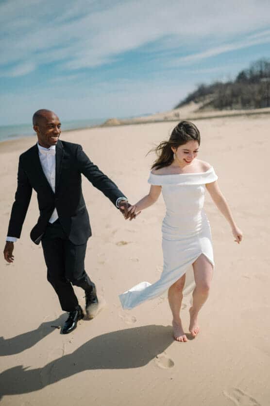 A smiling couple in wedding attire holds hands and walks barefoot on a sandy beach under a blue sky. The bride wears a white off-shoulder dress with a slit, and the groom wears a black suit. by Justin Salem Meyer
