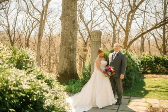 A bride in a white dress and a groom in a gray suit stand smiling outdoors, surrounded by greenery and tall trees with bare branches, holding a bouquet of flowers on a sunny day. by Justin Salem Meyer