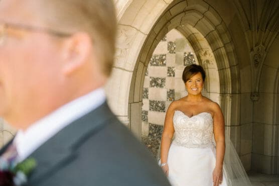 A smiling bride in a strapless white gown stands under a stone archway, while a man in a suit, blurred in the foreground, faces away from her. by Justin Salem Meyer