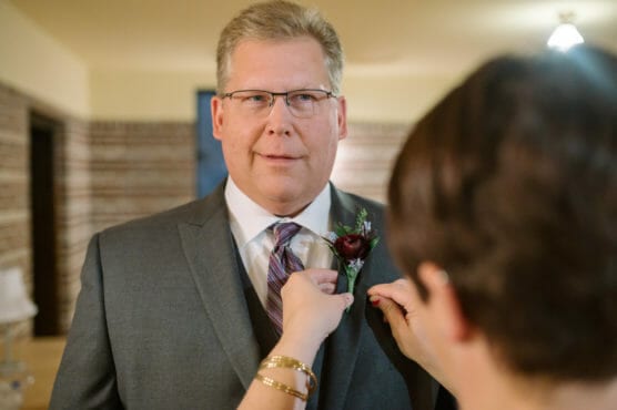 A man in a gray suit and striped tie stands indoors while someone pins a boutonniere with a dark red flower to his lapel. The man looks ahead with a slight smile in a warmly lit room. by Justin Salem Meyer