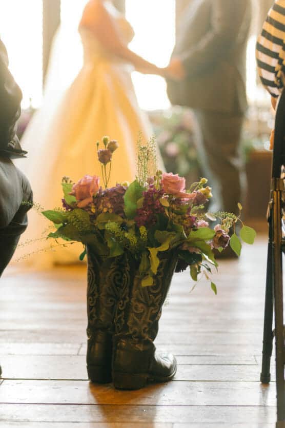 A pair of black cowboy boots filled with a colorful flower arrangement sits on a wooden floor. In the blurred background, a bride and groom stand holding hands during their wedding ceremony. by Justin Salem Meyer