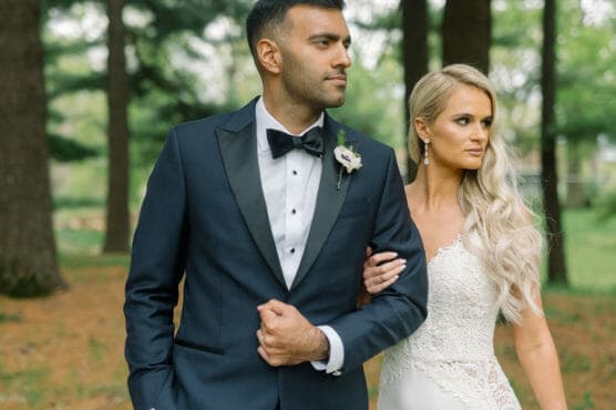 A groom in a dark tuxedo and a bride in a white lace dress stand arm in arm outdoors, looking into the distance with serious expressions. Tall trees and greenery are visible in the background. by Justin Salem Meyer