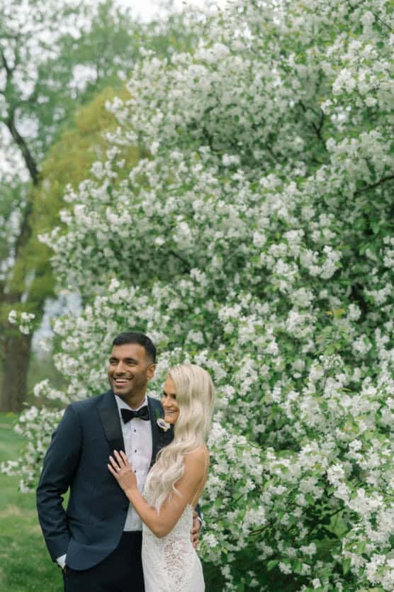 A bride and groom stand smiling together in front of a lush, blooming tree with white flowers. The groom wears a navy tuxedo, and the bride wears a white lace dress with long, wavy blonde hair. by Justin Salem Meyer
