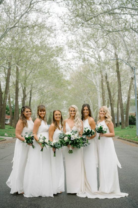 Six women in white dresses stand together outdoors on a tree-lined street, holding bouquets of white flowers and greenery, smiling at the camera. The scene is bright and serene, with spring foliage overhead. by Justin Salem Meyer