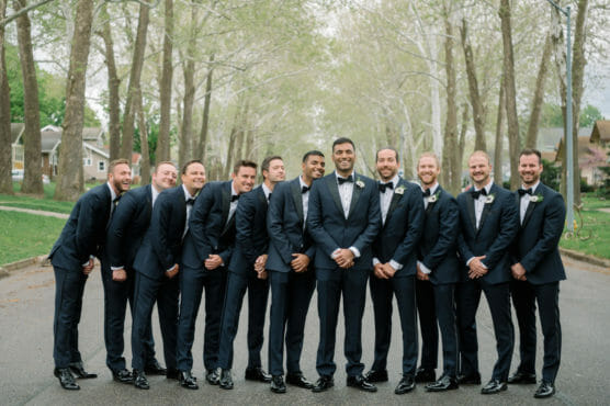 A group of twelve men dressed in matching dark suits, white shirts, and bow ties stand smiling together in the middle of a tree-lined street, posing for a formal photo on a cloudy day. by Justin Salem Meyer