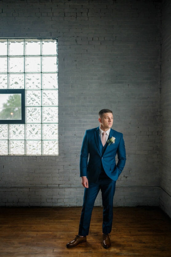 A man in a blue suit with a boutonniere stands on a wooden floor near a large glass-brick window, looking to his left against a gray brick wall. Natural light illuminates the scene. by Justin Salem Meyer