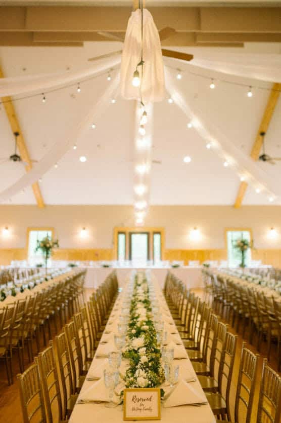 A decorated banquet hall with long tables set for an event, white tablecloths, gold chairs, floral centerpieces, hanging string lights, and draped white fabric on the ceiling. A Reserved sign is on the nearest table. by Justin Salem Meyer