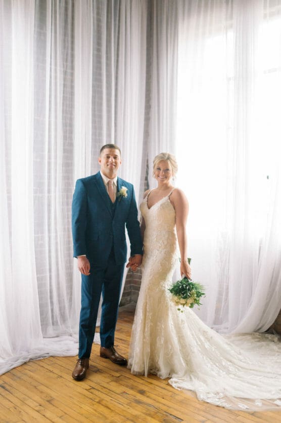 A bride in a lace wedding gown holding a bouquet stands beside a groom in a blue suit and pink tie. They are holding hands and posing in front of tall, sheer white curtains on a wooden floor. by Justin Salem Meyer