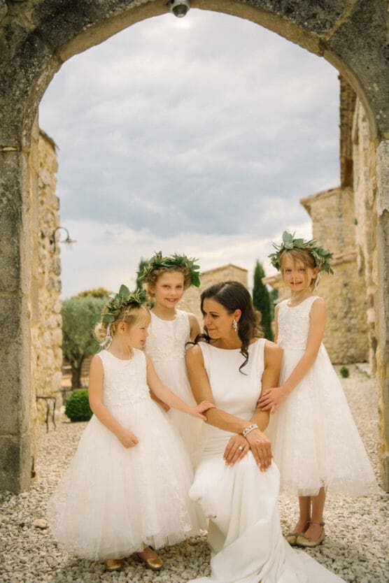 A bride in a white dress kneels outside under a stone arch, smiling at three young flower girls in white dresses and flower crowns, all posing happily together. by Justin Salem Meyer