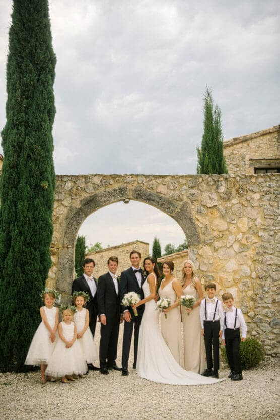 A wedding party poses outdoors by a stone archway and tall cypress trees. The group includes the bride, groom, bridesmaids in light dresses, groomsmen in suits, and four children in formal attire, all smiling at the camera. by Justin Salem Meyer