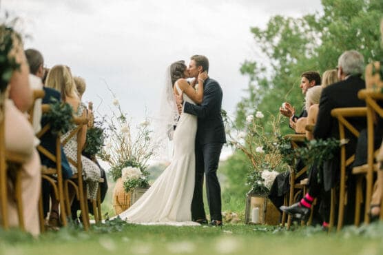 A bride and groom share a kiss at an outdoor wedding ceremony, surrounded by seated guests on both sides, with greenery and floral decorations in the background. by Justin Salem Meyer