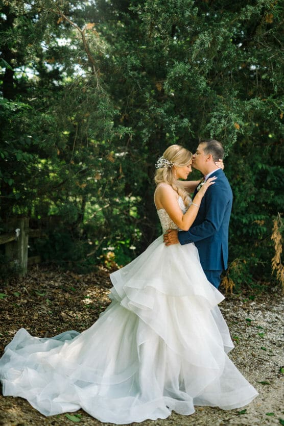 A bride in a white, layered gown and a groom in a blue suit lovingly embrace outdoors, surrounded by trees and greenery, with sunlight filtering through the branches. by Justin Salem Meyer