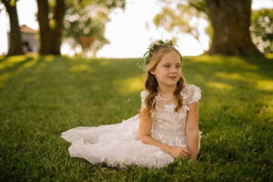 A young girl in a white dress with floral details and a flower crown sits on green grass in a sunlit park, looking to the side with a gentle expression. Trees and soft sunlight fill the background. by Justin Salem Meyer