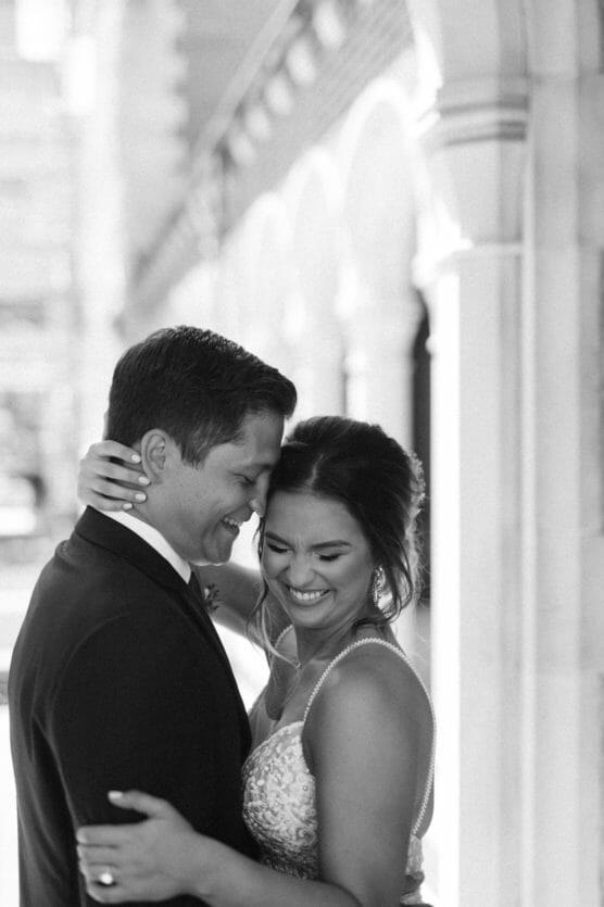 A joyful bride and groom embrace closely, smiling and touching foreheads, standing together in formal wedding attire near an elegant arched walkway. The photo is in black and white. by Justin Salem Meyer