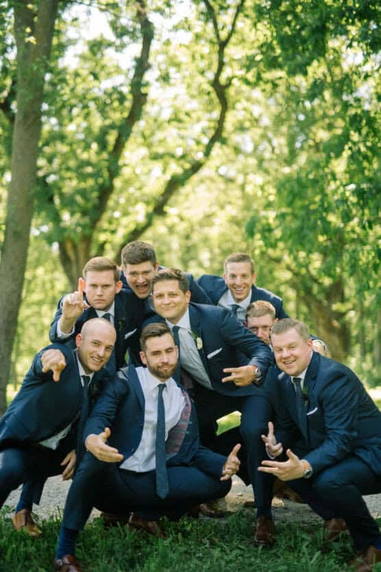 Eight men in navy suits pose playfully in a wooded outdoor area, crouching and making hand gestures while smiling and laughing. Sunlight filters through the trees in the background. by Justin Salem Meyer