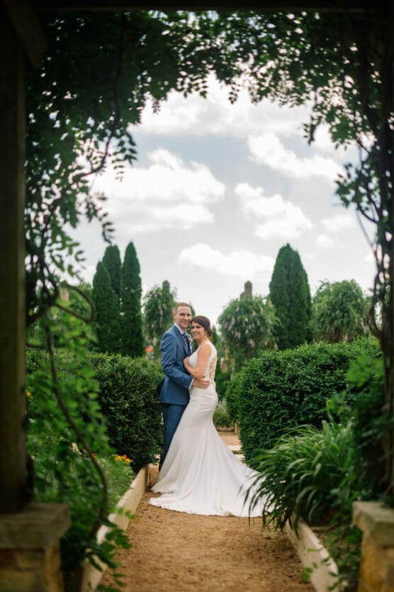 A bride and groom stand together on a garden path, surrounded by lush greenery and hedges, framed by an archway, with a bright sky and fluffy clouds overhead. by Justin Salem Meyer